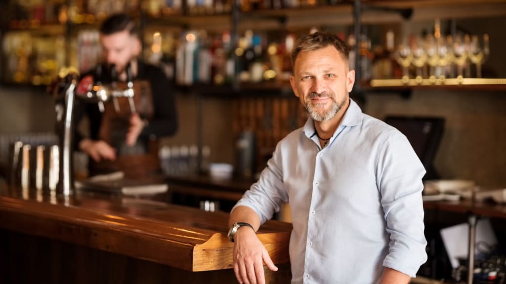 A man in a white shirt stands at a wooden bar in California, smiling as the bartender works in the background, likely discussing a liquor license transfer.